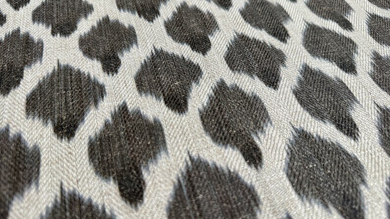 May include: Close-up of a white fabric with a black ikat pattern. The fabric has a subtle herringbone weave.