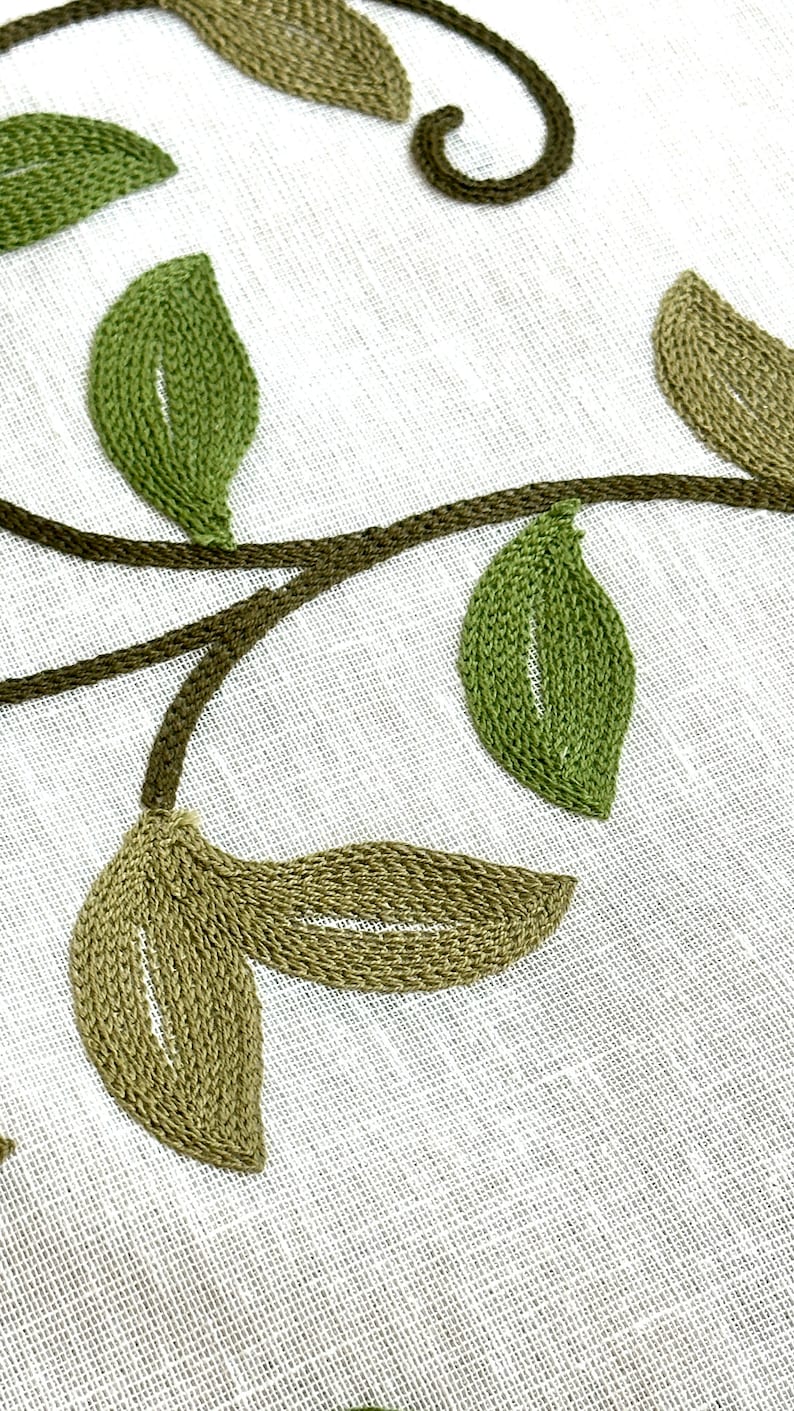 May include: Embroidered green leaves and vines on a white linen fabric background.