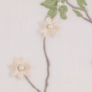 May include: White fabric with embroidered floral design. The flowers are white with a pearl center and the leaves are green.