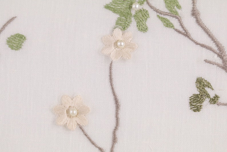 May include: White fabric with embroidered floral design. The flowers are white with a pearl center and the leaves are green.