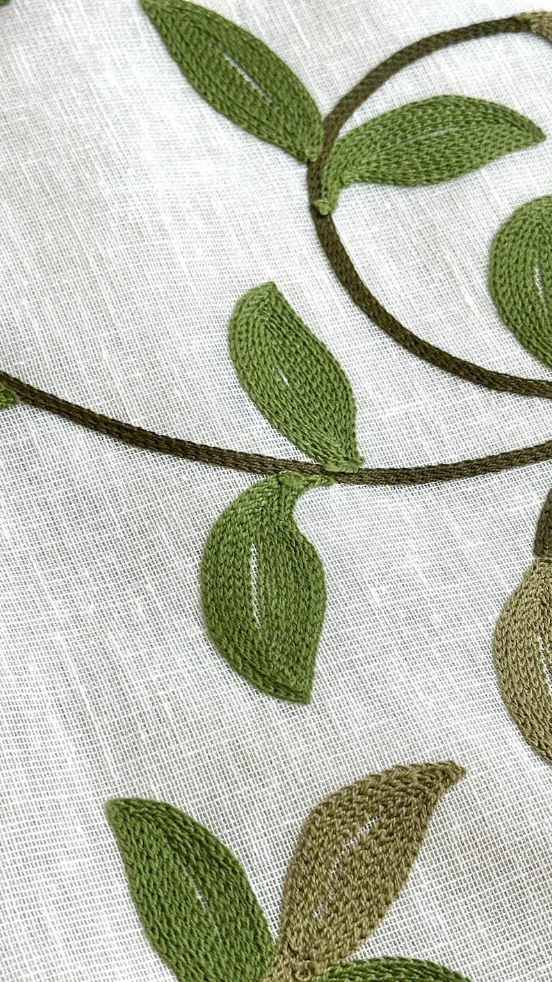 May include: Close-up of a white fabric with green embroidered leaves and vines. The leaves are stitched with a detailed, textured pattern.