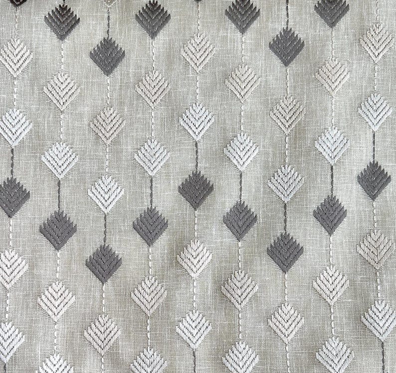 May include: Close-up of a fabric with a repeating pattern of embroidered diamond shapes. The diamonds alternate between light gray and dark gray, connected by vertical lines of stitching on a light beige background. The fabric has a textured appearance.