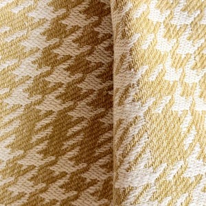 May include: A woven fabric featuring a repeating houndstooth pattern in mustard yellow and off-white. The textile displays a dense, textured weave with visible interlacing threads, creating a consistent geometric design across the surface.