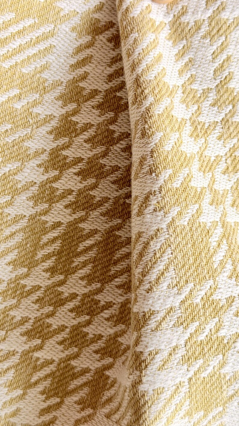 May include: A woven fabric featuring a repeating houndstooth pattern in mustard yellow and off-white. The textile displays a dense, textured weave with visible interlacing threads, creating a consistent geometric design across the surface.