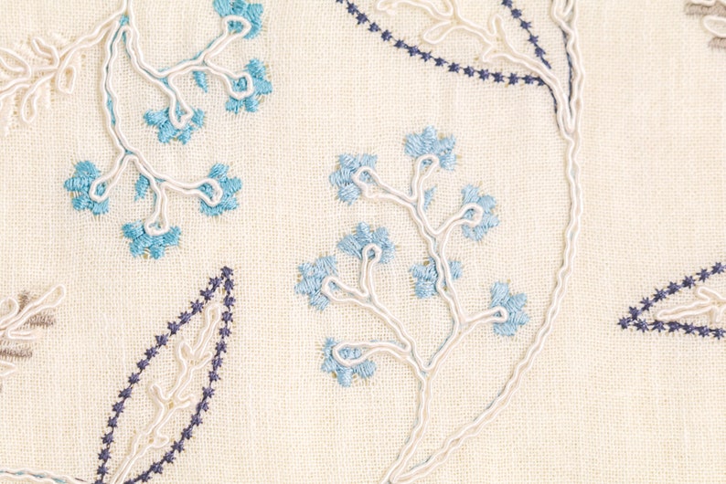 Puede incluir: Primer plano de una tela color crema con patrones florales y de hojas bordados. El bordado presenta hilo azul claro y azul oscuro, con contornos de cord&oacute;n blanco. El dise&ntilde;o incluye flores y hojas estilizadas.