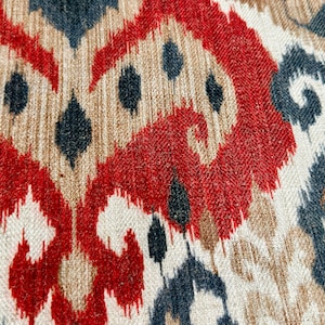 May include: Close-up of a fabric with a red, brown, and blue ikat pattern. The fabric has a woven texture and a repeating design.