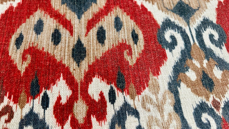 May include: Close-up of a fabric with a red, brown, and blue ikat pattern. The fabric has a woven texture and a repeating design.