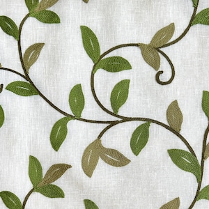 May include: A white fabric with an embroidered design of green leaves and vines. The leaves are in various shades of green and are arranged in a flowing pattern.