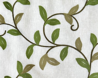 May include: A white fabric with an embroidered design of green leaves and vines. The leaves are in various shades of green and are arranged in a flowing pattern.
