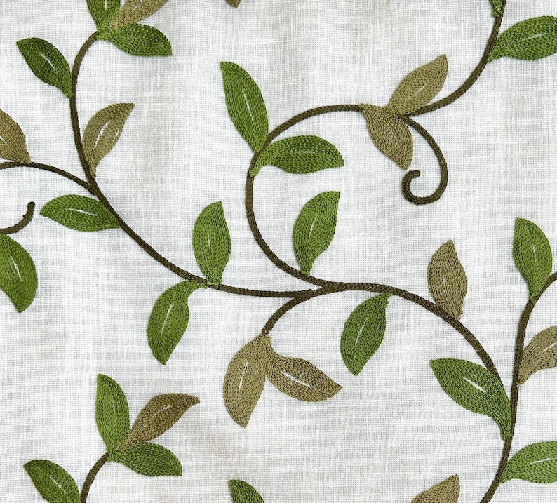 May include: A white fabric with an embroidered design of green leaves and vines. The leaves are in various shades of green and are arranged in a flowing pattern.