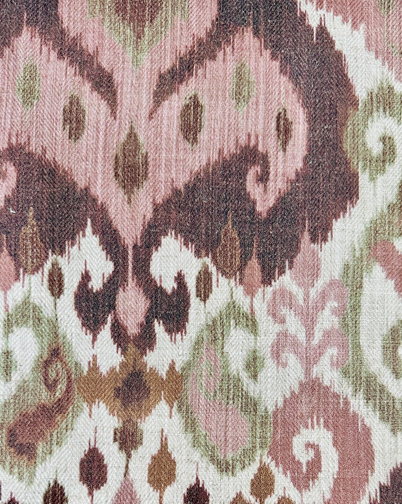 May include: Close-up of a woven fabric with an ikat pattern. The design features repeating abstract shapes in shades of brown, pink, green, and off-white. The texture of the fabric is visible, adding depth to the design.