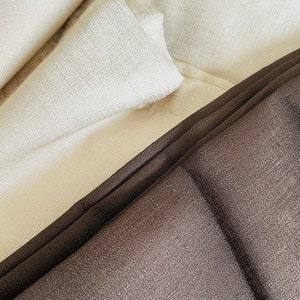 May include: Close-up of folded fabric in shades of cream and dark brown. The fabric has a textured appearance, with visible woven details. The folds create shadows and highlights, adding depth to the image. The overall aesthetic is simple and elegant.