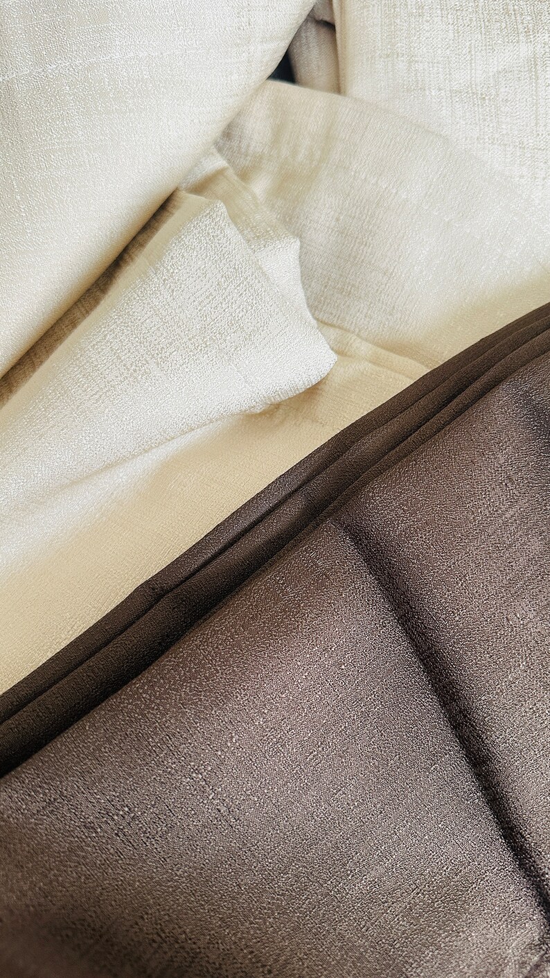 May include: Close-up of folded fabric in shades of cream and dark brown. The fabric has a textured appearance, with visible woven details. The folds create shadows and highlights, adding depth to the image. The overall aesthetic is simple and elegant.
