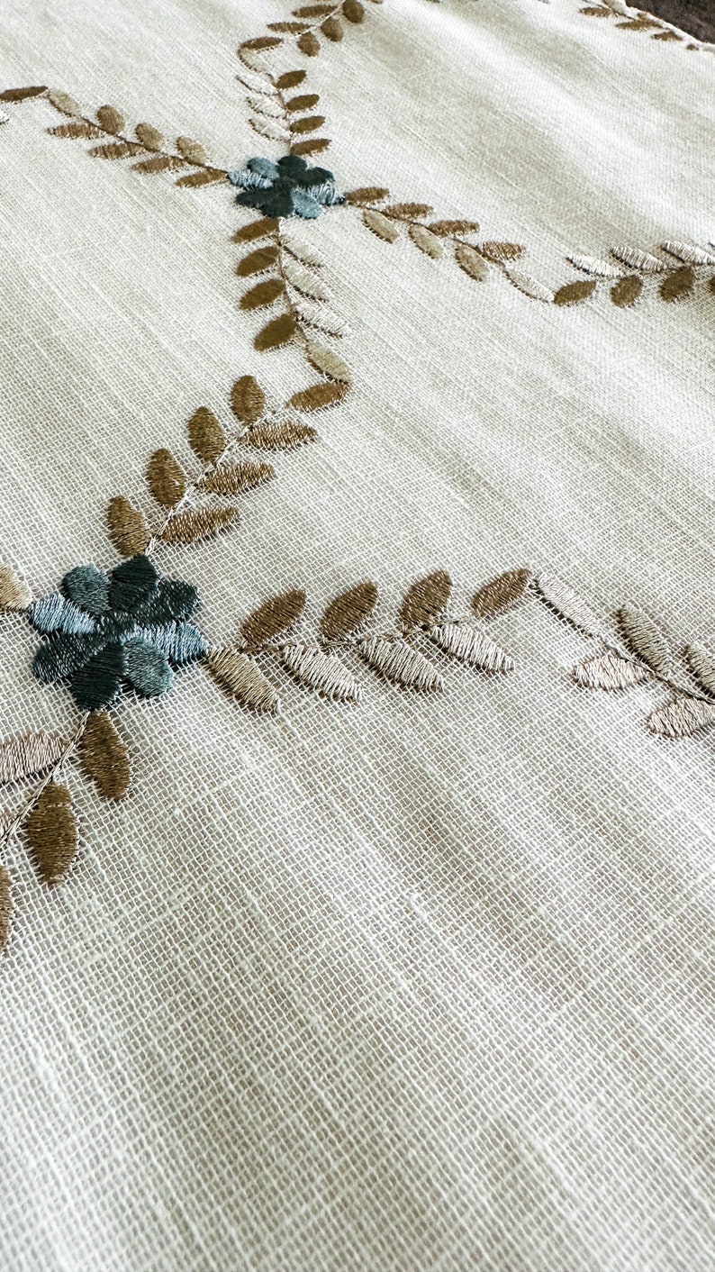 Puede incluir: Tela blanca con hojas marrones y flores verdes bordadas.
