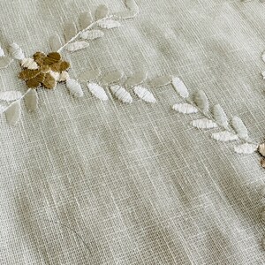 May include: Close-up of a beige fabric with embroidered white leaves and brown flowers.
