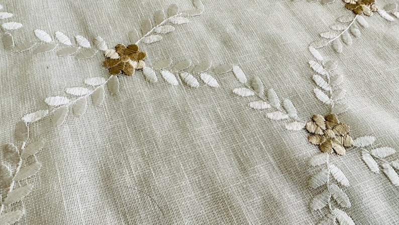 May include: Close-up of a beige fabric with embroidered white leaves and brown flowers.