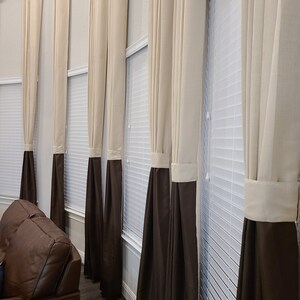 May include: A series of tall, elegant curtains in a room. The curtains are a combination of beige and dark brown, with a band of beige at the top and a dark brown bottom. The curtains hang in front of white blinds, creating a layered effect.