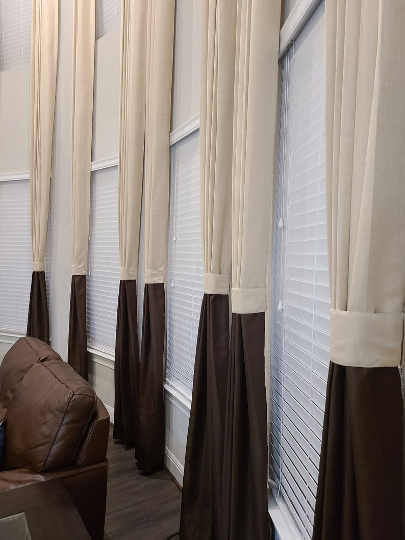 May include: A series of tall, elegant curtains in a room. The curtains are a combination of beige and dark brown, with a band of beige at the top and a dark brown bottom. The curtains hang in front of white blinds, creating a layered effect.