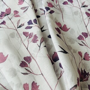 May include: White fabric with a floral pattern featuring pink and purple flowers and stems. The fabric is folded and draped, showing the texture and design.