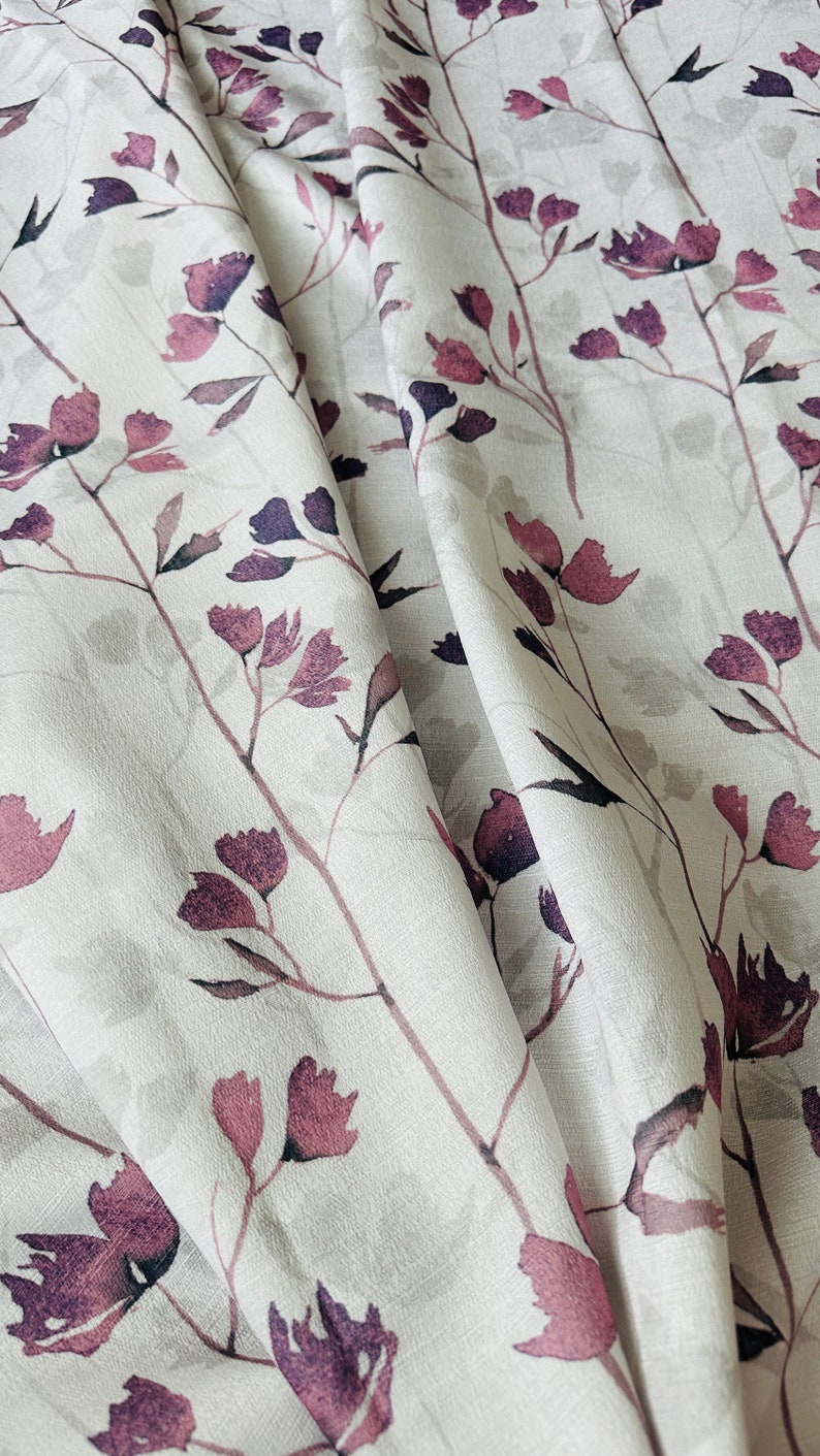 May include: White fabric with a floral pattern featuring pink and purple flowers and stems. The fabric is folded and draped, showing the texture and design.