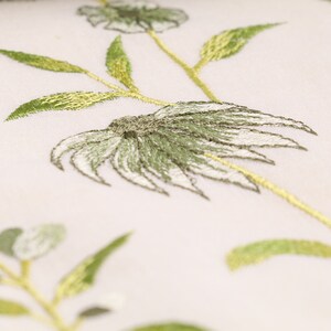 May include: Close-up of a white fabric with green embroidered floral designs. The flowers have a delicate, layered look with a white center and green petals.