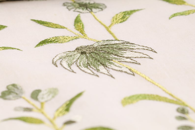 May include: Close-up of a white fabric with green embroidered floral designs. The flowers have a delicate, layered look with a white center and green petals.