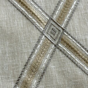 Pu&ograve; includere: Primo piano di un tessuto beige con due bande di ricamo beige e grigio che si incrociano. Il ricamo &egrave; cucito in linea retta con un motivo decorativo.