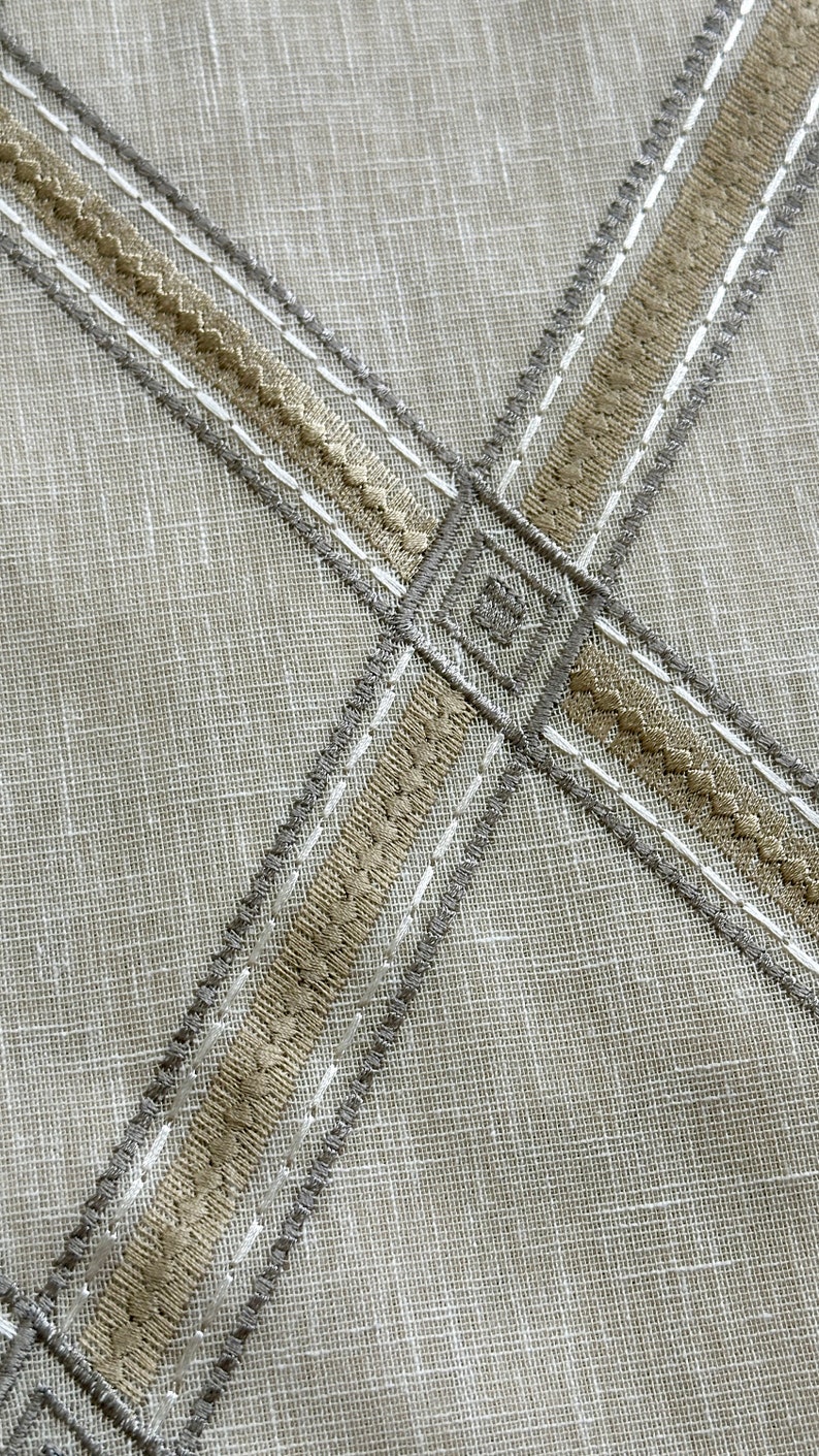 Pu&ograve; includere: Primo piano di un tessuto beige con due bande di ricamo beige e grigio che si incrociano. Il ricamo &egrave; cucito in linea retta con un motivo decorativo.
