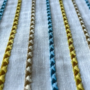 May include: Close-up of a white fabric with embroidered stripes in yellow, blue, and beige.