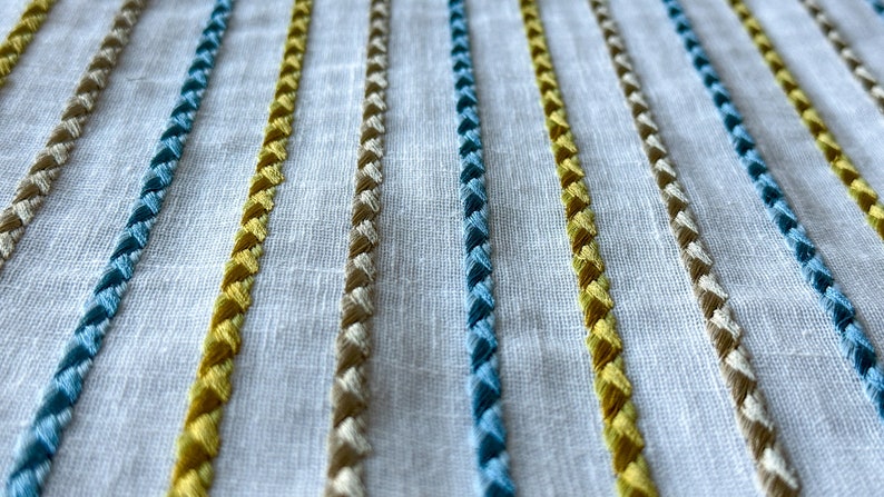 May include: Close-up of a white fabric with embroidered stripes in yellow, blue, and beige.