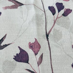 May include: A close-up of a white fabric with a subtle textured pattern. The fabric features a delicate floral design with purple and pink flowers and leaves.