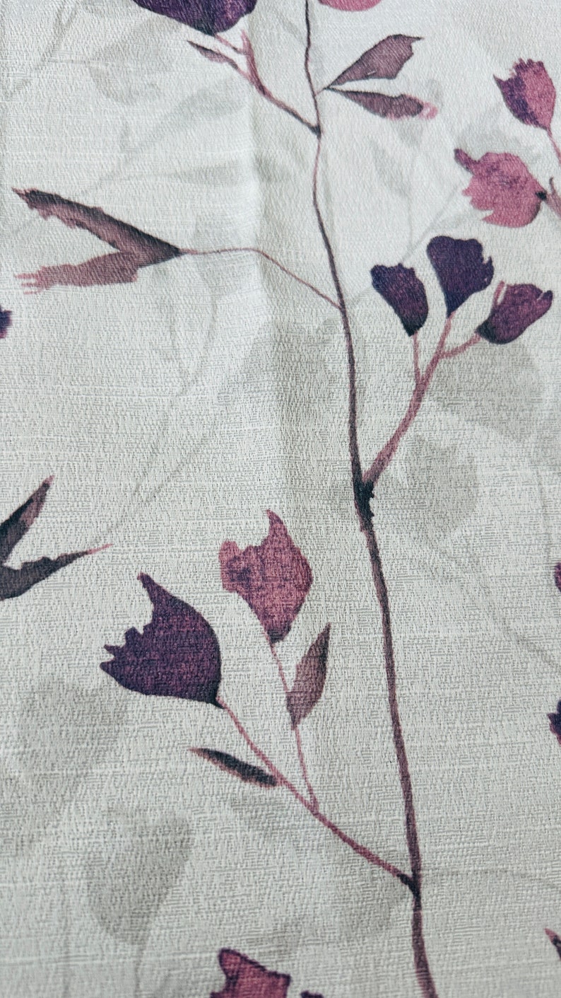 May include: A close-up of a white fabric with a subtle textured pattern. The fabric features a delicate floral design with purple and pink flowers and leaves.