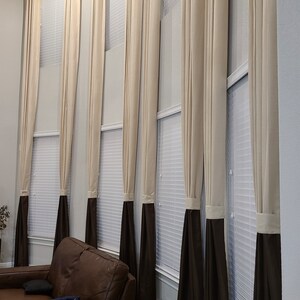 May include: Tall, cream-colored curtains with a dark brown bottom section hang in front of a window. The curtains are pleated and reach the floor. A brown leather sofa is in the foreground.