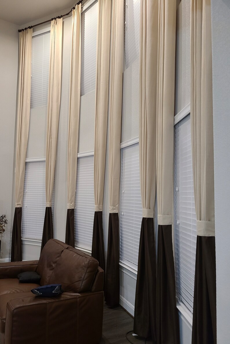 May include: Tall, cream-colored curtains with a dark brown bottom section hang in front of a window. The curtains are pleated and reach the floor. A brown leather sofa is in the foreground.