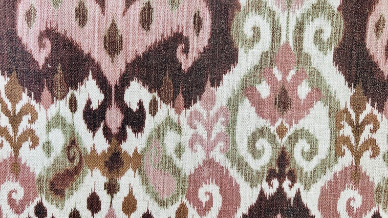 May include: Textile with an ikat pattern in shades of brown, pink, green, and white. The design features stylized floral and geometric shapes, creating a textured, woven appearance. The fabric has a vintage or bohemian aesthetic.