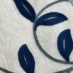 May include: Close-up of a fabric with an embroidered design of blue and gray leaves. The fabric has a textured, linen-like appearance.