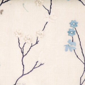 May include: White fabric with a floral pattern featuring blue and white flowers and pearl accents. The fabric has a subtle texture and a delicate design.