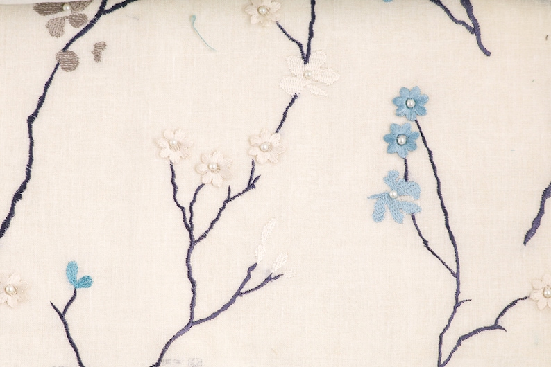May include: White fabric with a floral pattern featuring blue and white flowers and pearl accents. The fabric has a subtle texture and a delicate design.