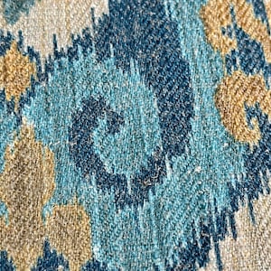 May include: Close-up of a fabric with a blue, teal, and beige ikat pattern. The fabric has a woven texture and a subtle sheen.