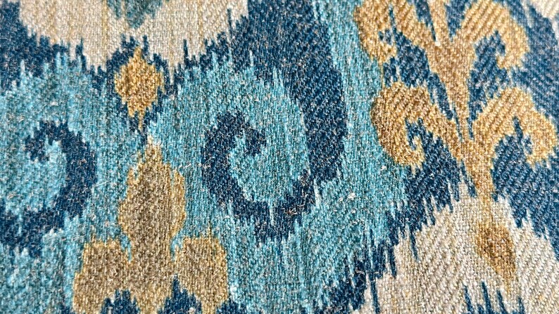 May include: Close-up of a fabric with a blue, teal, and beige ikat pattern. The fabric has a woven texture and a subtle sheen.