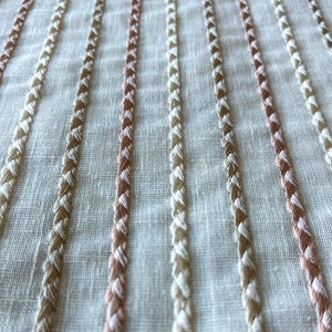 Puede incluir: Tela blanca con filas verticales de costuras bordadas en beige y rosa.