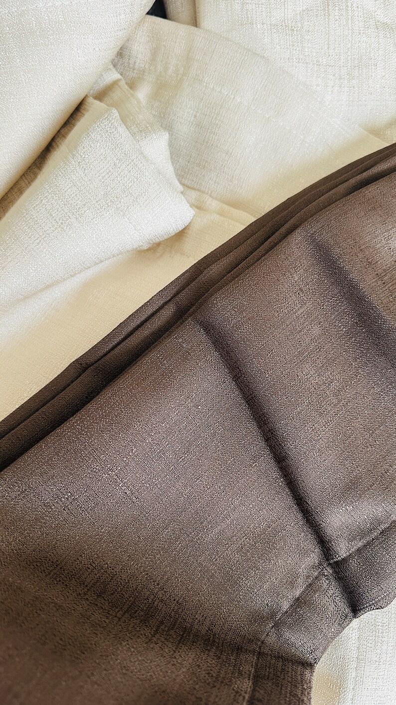 May include: Close-up of folded fabric in shades of brown and off-white. The brown fabric has a textured appearance, while the off-white fabric appears smooth. The image highlights the different textures and colors of the textiles.
