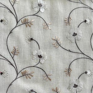 May include: A beige fabric with an embroidered floral pattern. The flowers are white with brown centres and are arranged in a repeating pattern on the fabric.