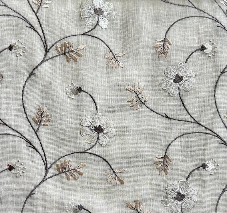 May include: A beige fabric with an embroidered floral pattern. The flowers are white with brown centres and are arranged in a repeating pattern on the fabric.