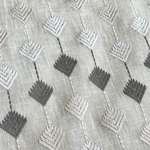 May include: Close-up of a textured fabric with a repeating pattern of diamond-shaped embroidered designs. The diamonds alternate between light gray and dark gray, connected by thin vertical lines on a light beige background.