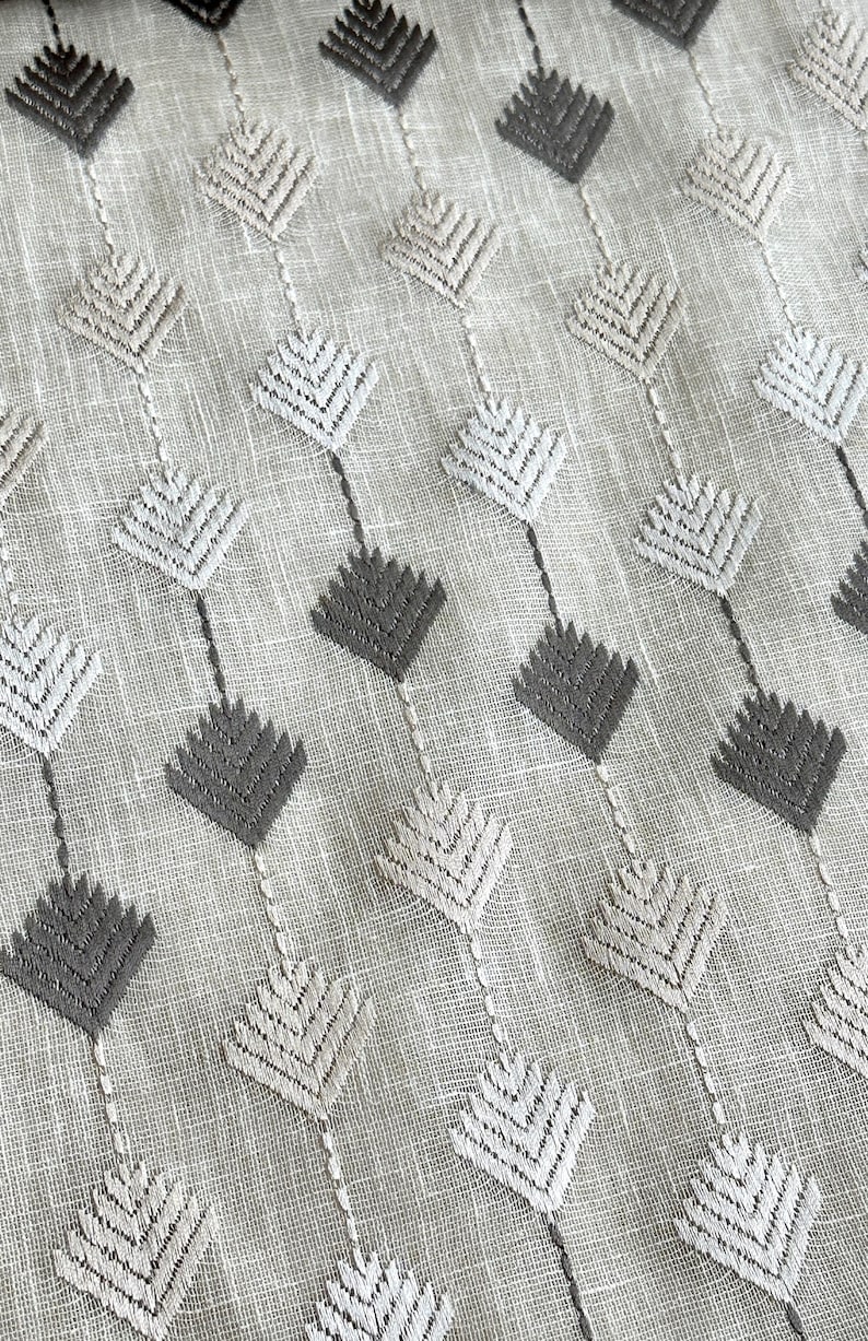 May include: Close-up of a textured fabric with a repeating pattern of diamond-shaped embroidered designs. The diamonds alternate between light gray and dark gray, connected by thin vertical lines on a light beige background.