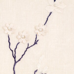 May include: White and blue embroidered floral design on a cream fabric background. The flowers are embellished with small pearl beads.