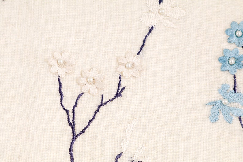 May include: White and blue embroidered floral design on a cream fabric background. The flowers are embellished with small pearl beads.
