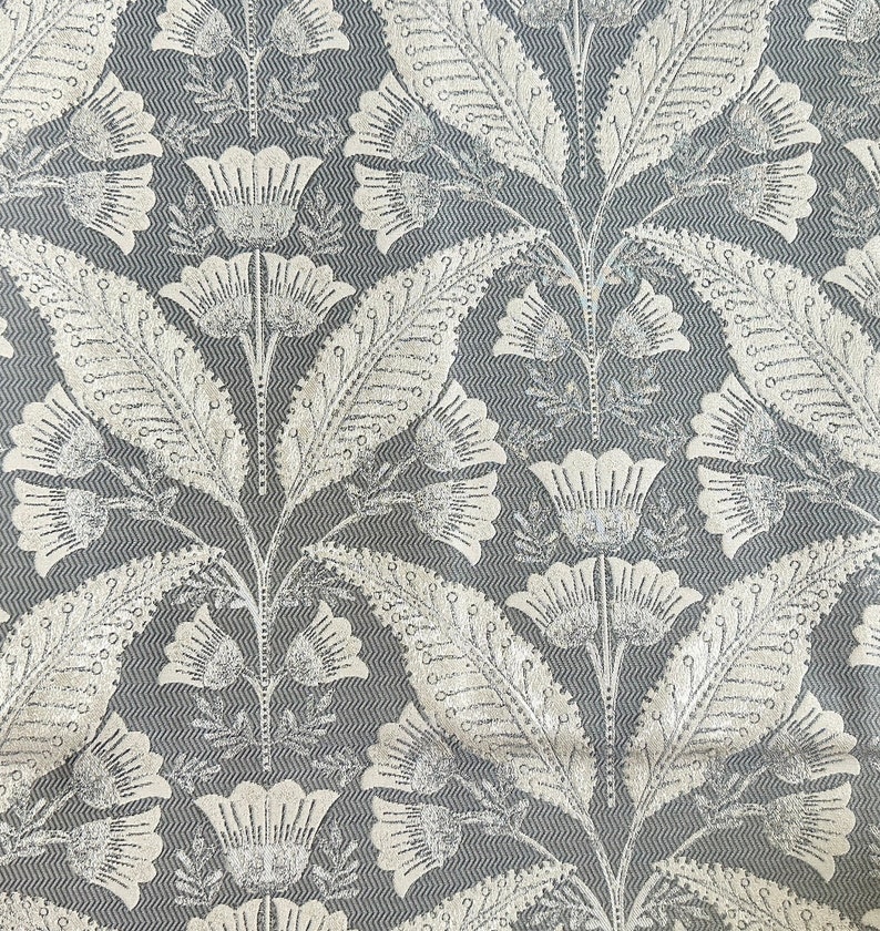 Pu&ograve; includere: Un tessuto grigio e bianco con un motivo ripetitivo di fiori e foglie stilizzati. Il tessuto ha una texture a spina di pesce sottile.
