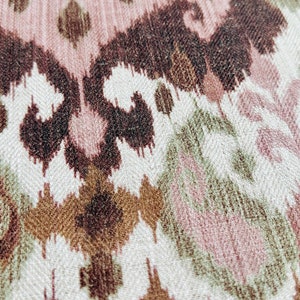 May include: Close-up of a woven fabric with an ikat pattern. The design features repeating shapes in shades of brown, pink, and green on a cream background. The fabric has a textured appearance.